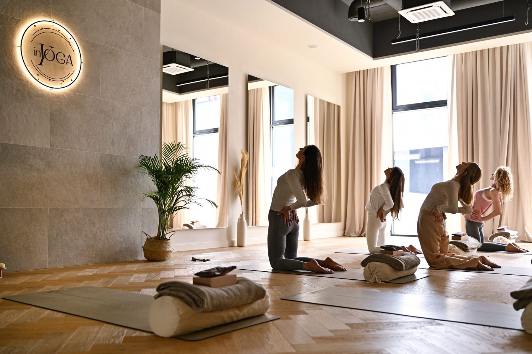Sala jogi inYOGA: kobiety w pozycji wielbłąda na matach, duże okna, podświetlone logo na ścianie